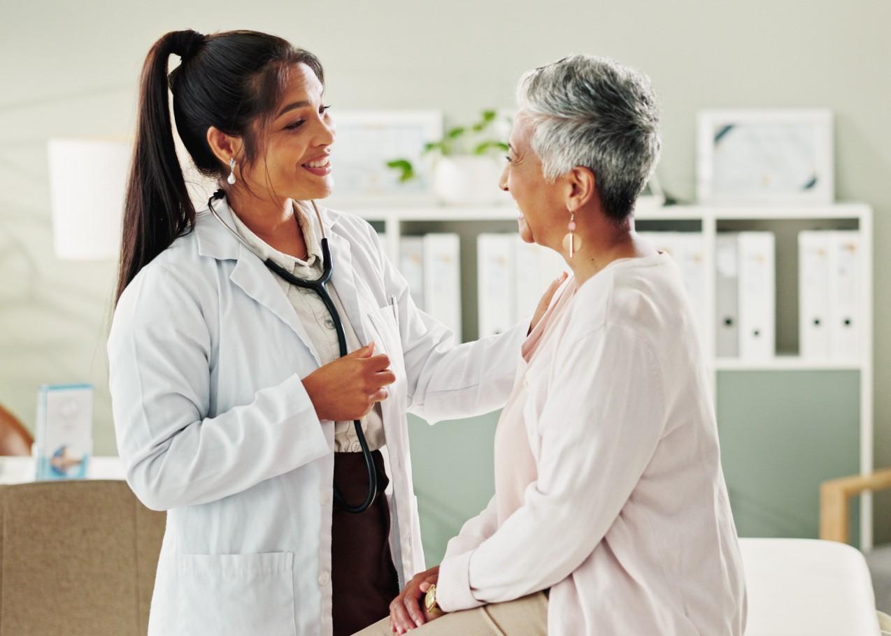 Smiling female doctor with a stethoscope reassuring an older woman in a medical office.