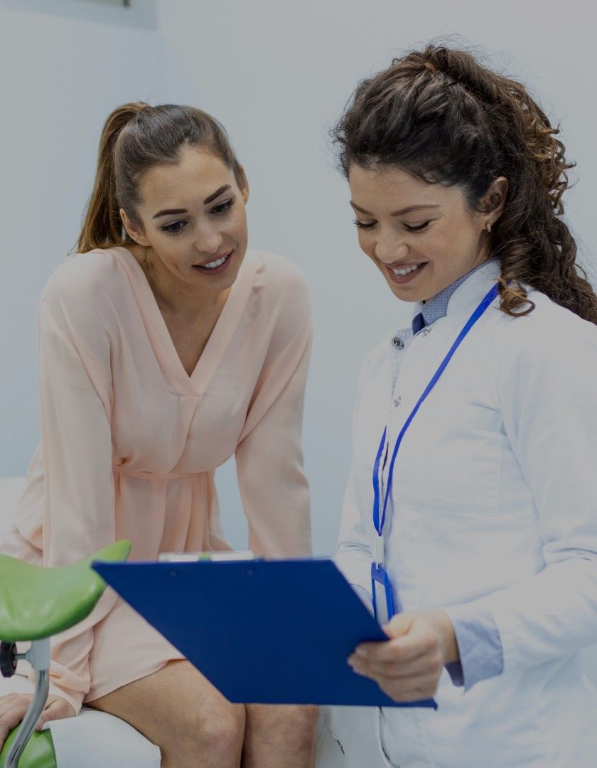 Female doctor in white coat showing medical clipboard to a seated female patient in a light pink dress.
