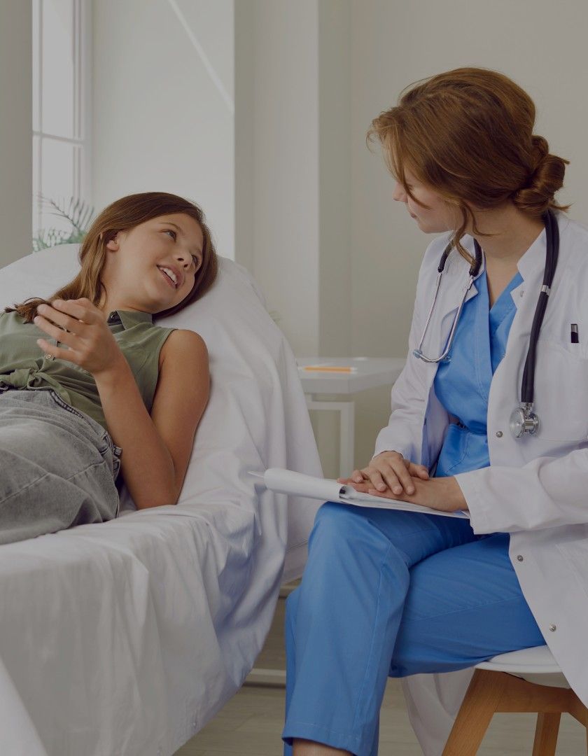 Female doctor in blue scrubs and white coat talking to a girl lying on a hospital bed in a bright room.