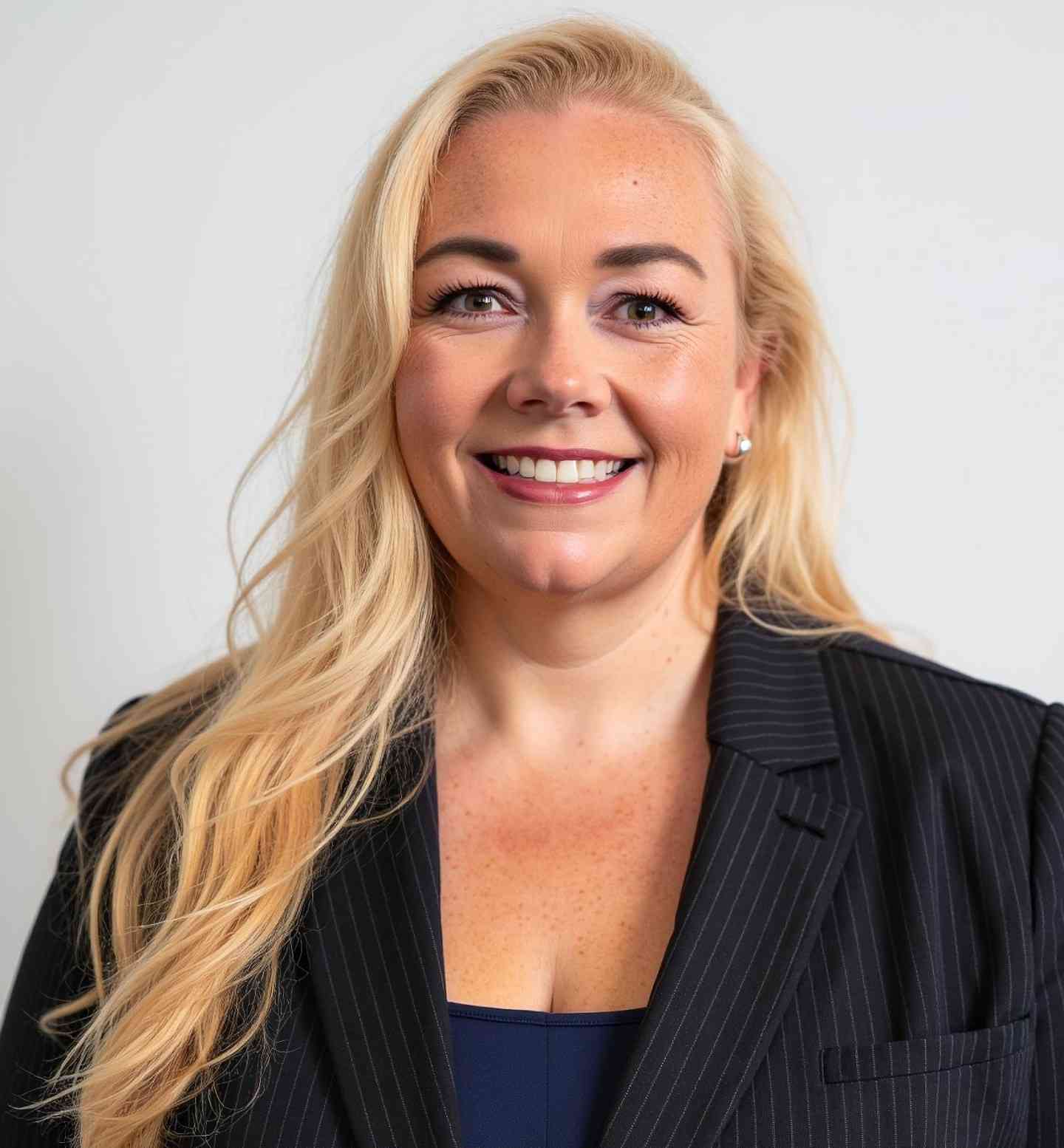 Smiling blonde woman with long hair wearing a black pinstripe blazer and a navy top against a light background.