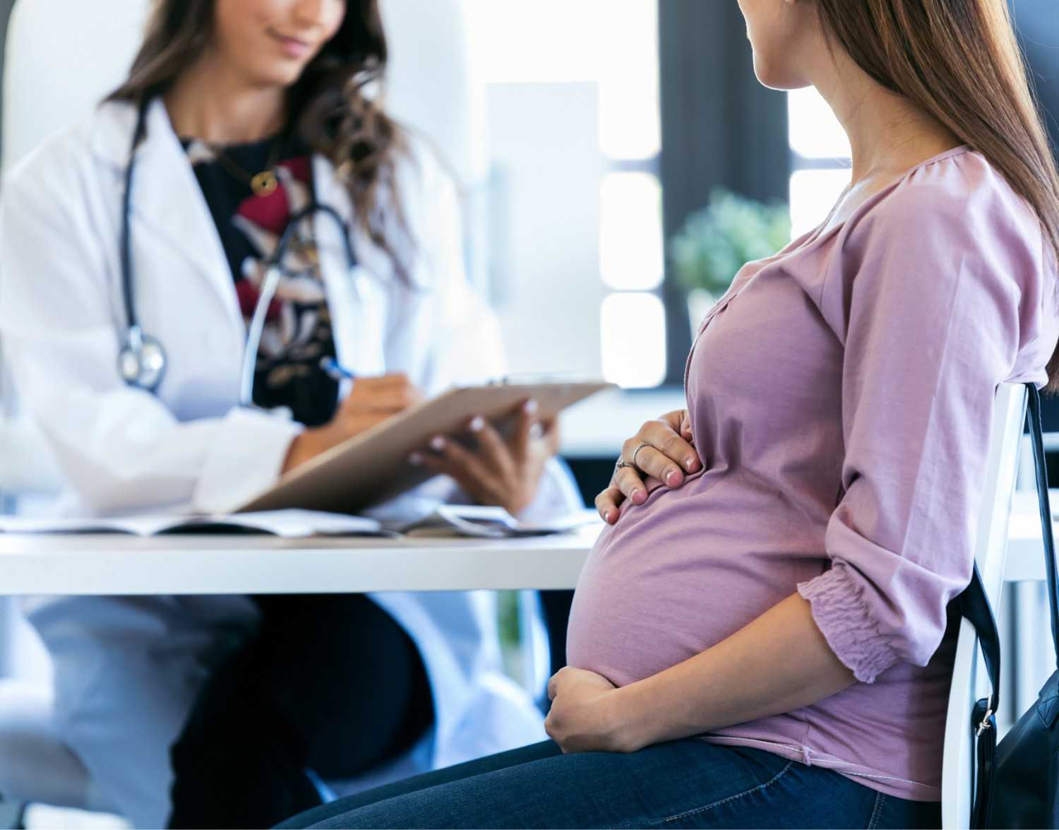 Pregnant woman in a pink top sitting and holding her belly during a medical consultation with a doctor.