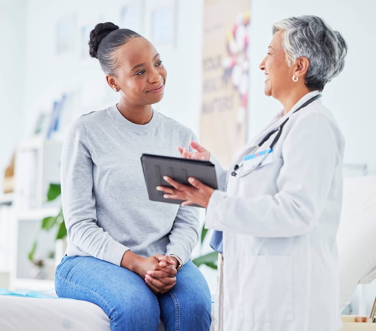 Smiling female doctor holding a tablet and talking with a young woman sitting on an exam table.