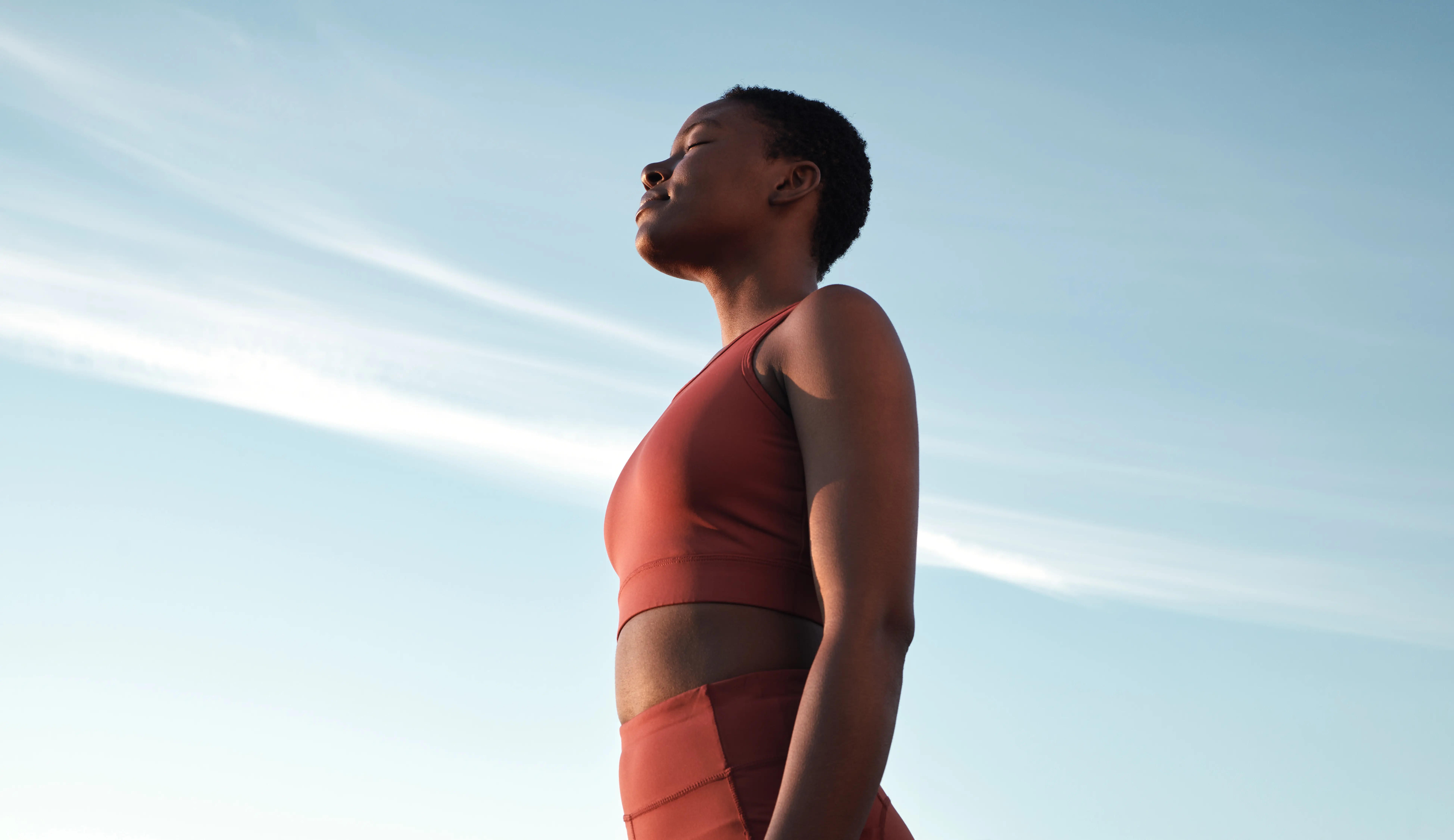 Woman with short curly hair wearing a red sports outfit standing peacefully against a blue sky with white clouds.