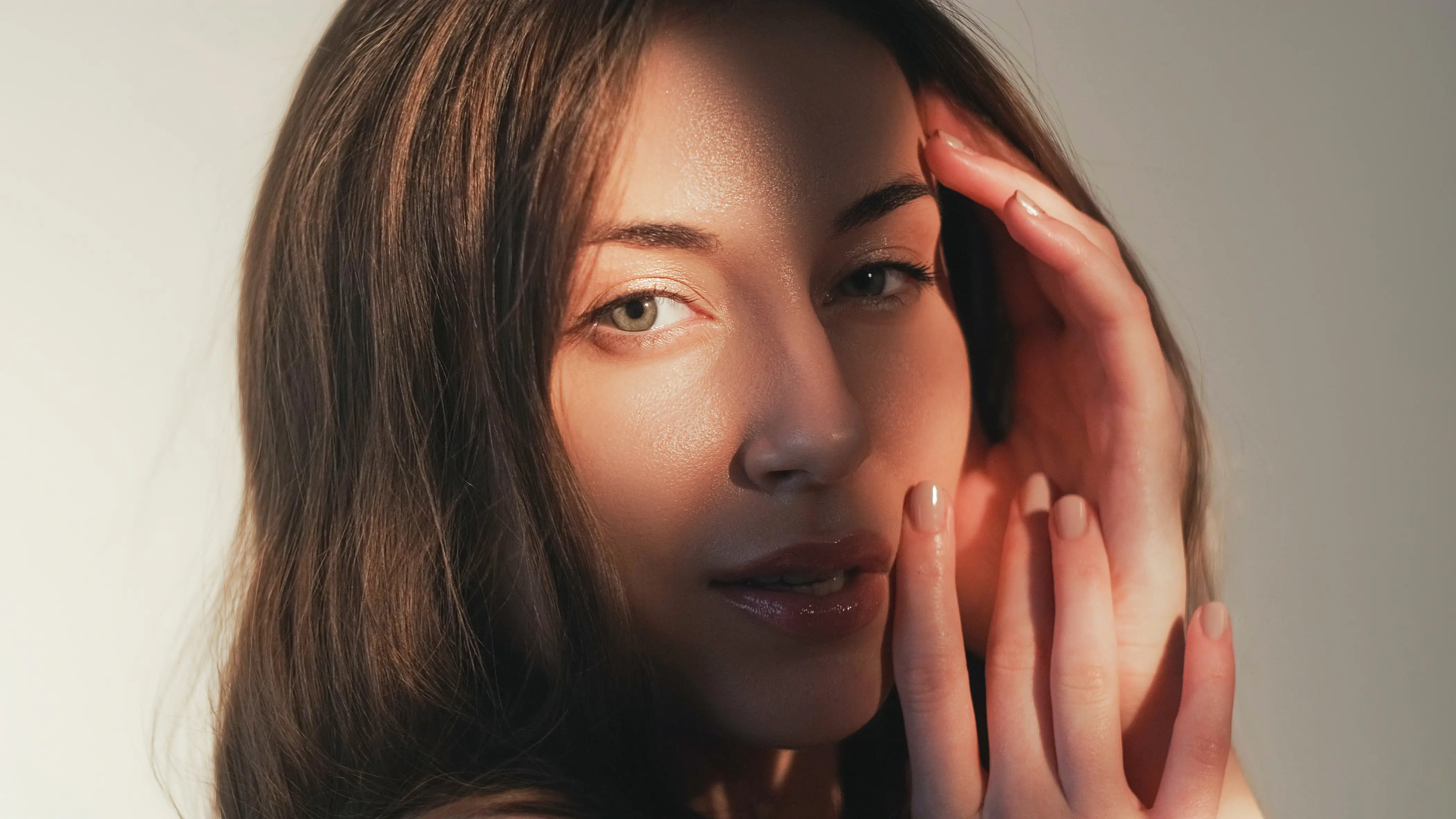Close-up of a woman with long brown hair gently touching her face with soft natural light and shadow.