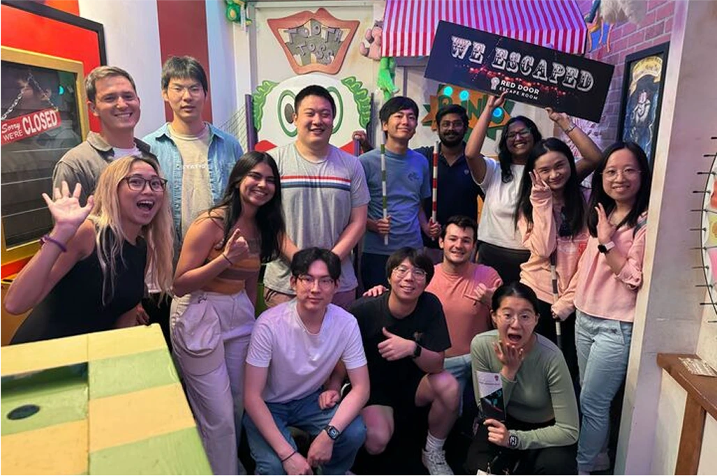 Large group of diverse young adults posing in an escape room, holding a "we escaped" sign.