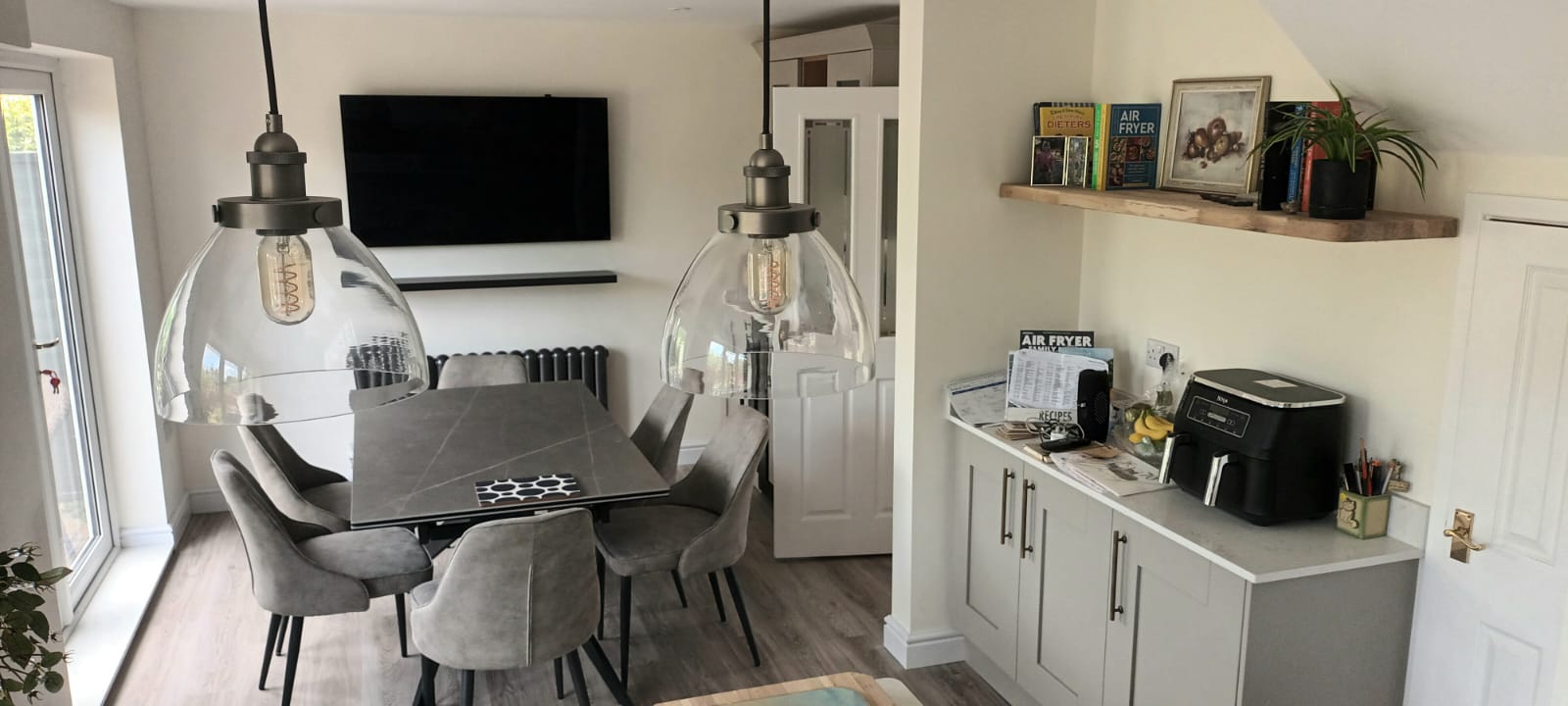 Modern dining room with grey chairs, glass pendant lights, wall-mounted TV, and fitted cabinetry after renovation.