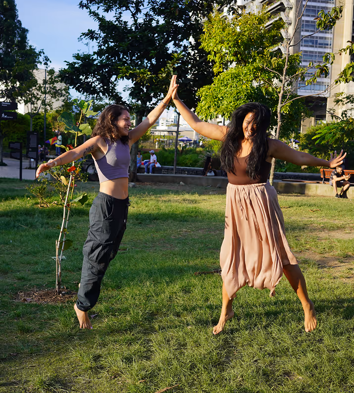 Two women barefoot, smiling and jumping in a grassy park while giving each other a high-five.