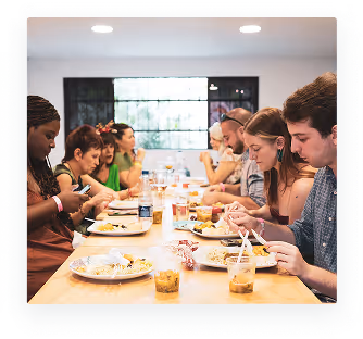 Group of diverse young adults seated at a long table enjoying a meal together indoors.