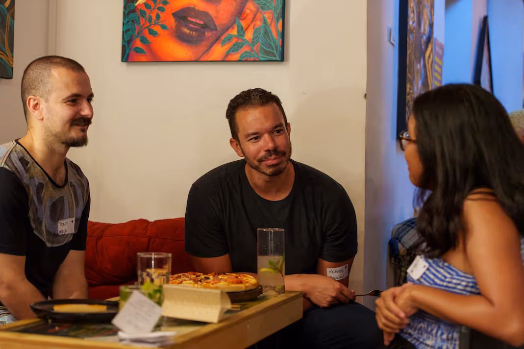 Three people seated around a table with a pizza and drinks, engaged in conversation in a cozy room with colorful artwork on the walls.