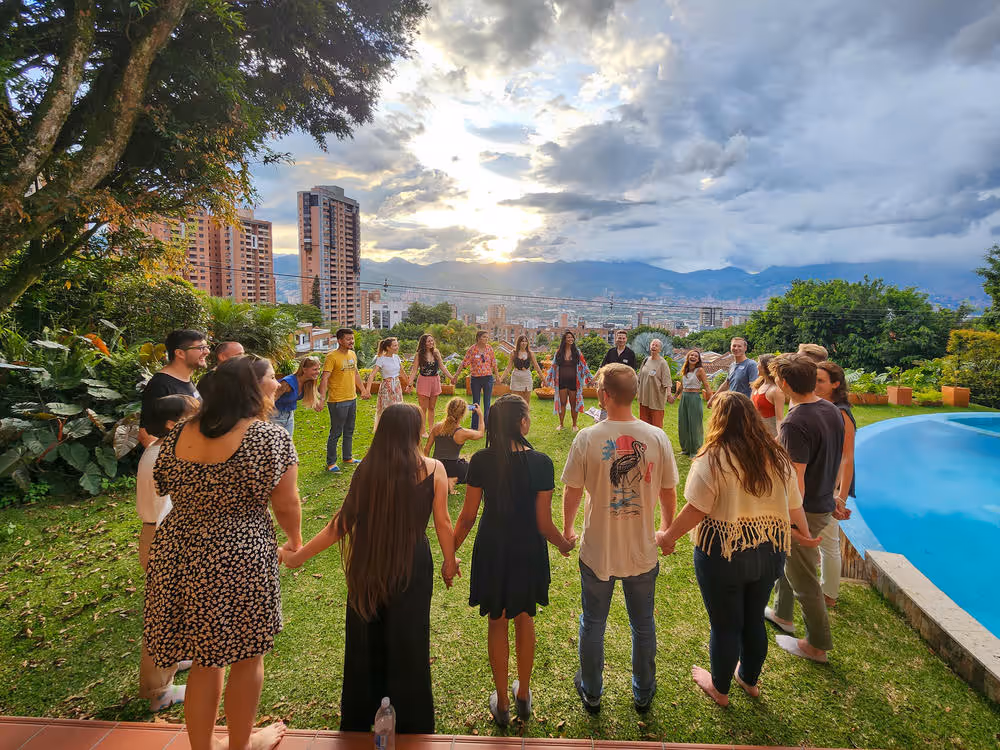 Group of people standing in a circle holding hands on a grassy lawn with a city and mountains in the background at sunset.