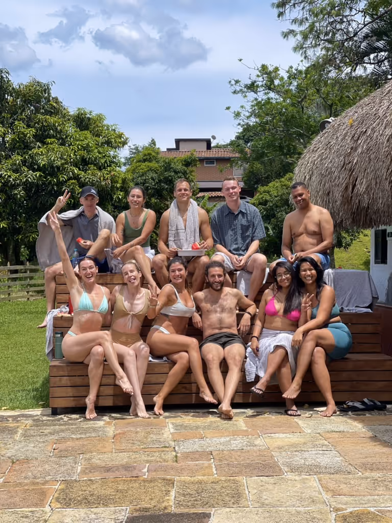Group of eleven people in swimwear sitting on wooden steps outdoors, smiling and posing under a partly cloudy sky with greenery in the background.