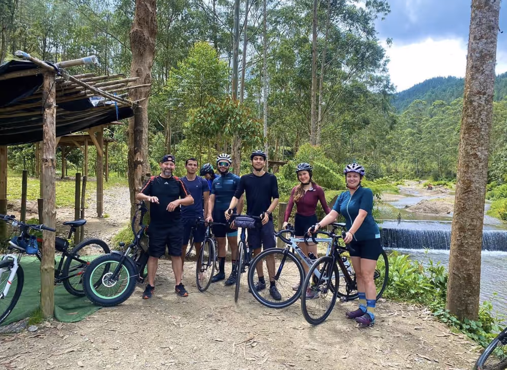 Group of six cyclists posing with their bikes on a dirt path near a small waterfall and lush green forest.