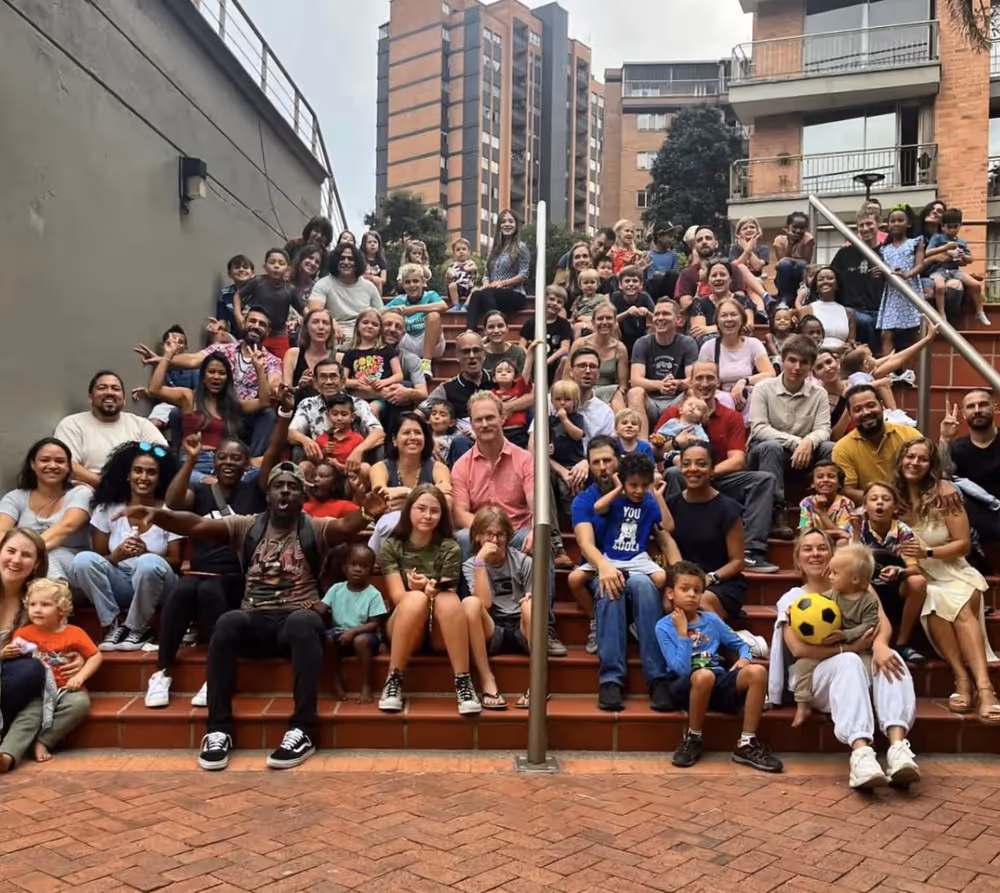 Large diverse group of adults and children sitting on outdoor steps with buildings in the background.