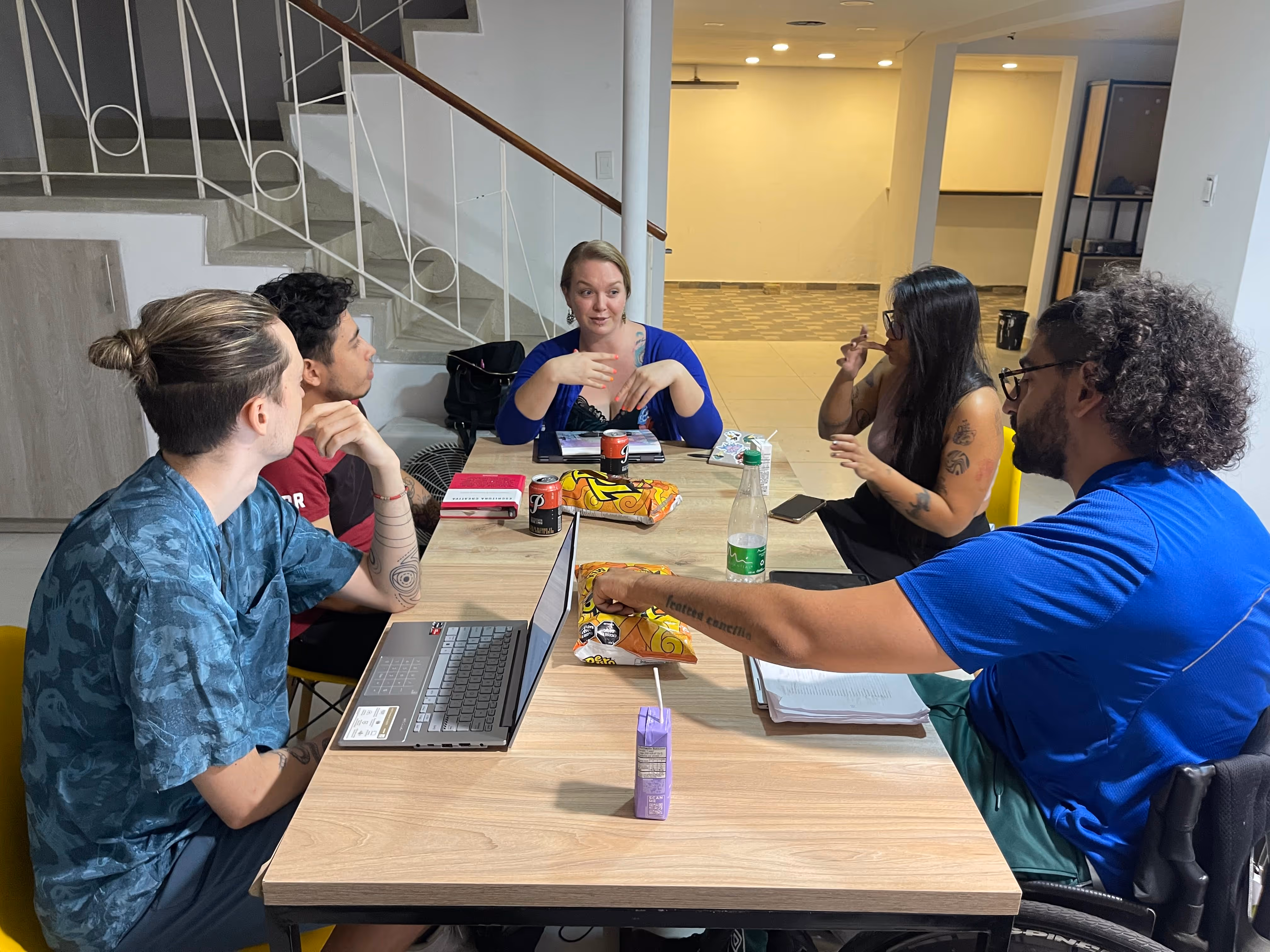 Five young adults sitting around a table in discussion, with laptops, notebooks, snacks, and drinks.