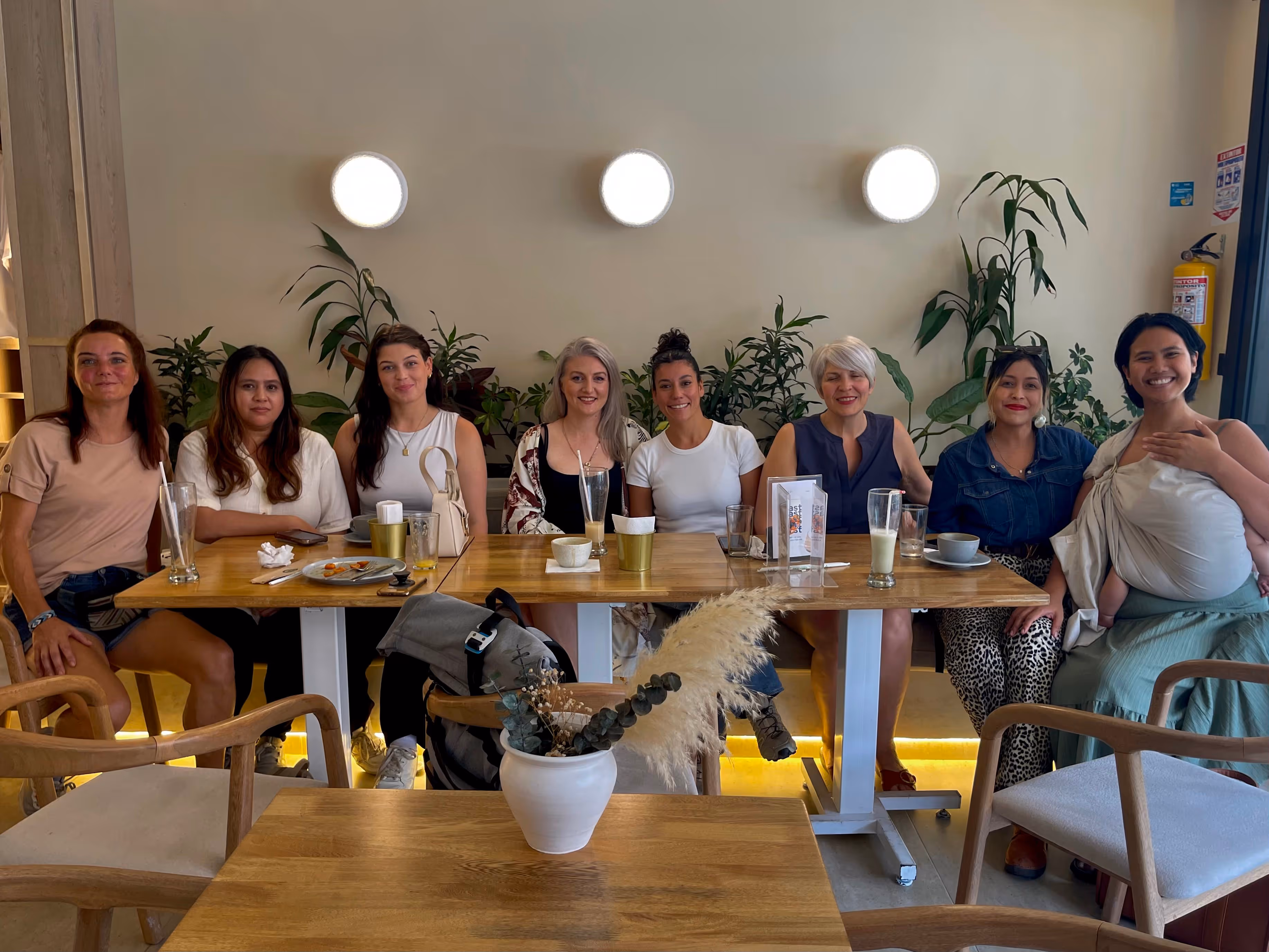 Group of eight diverse women sitting at a dining table in a cafe, with plants and three round wall lights behind them.