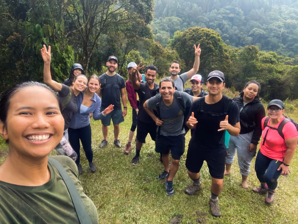 Group of smiling hikers posing outdoors on grassy terrain with a forested mountain background.