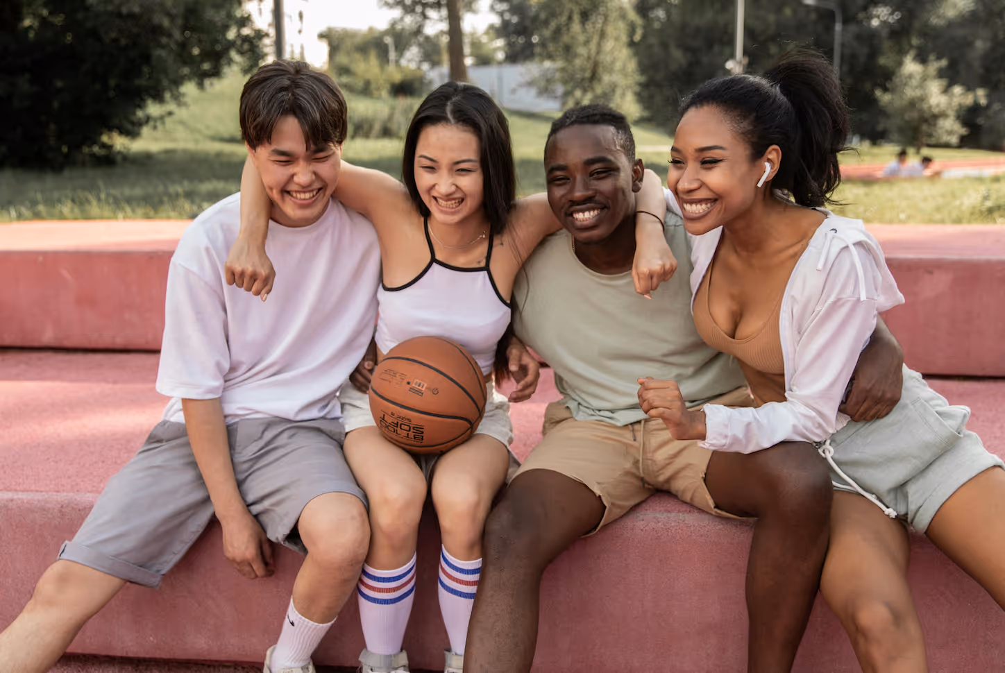 Four diverse young adults sitting closely on outdoor steps, smiling and holding a basketball.