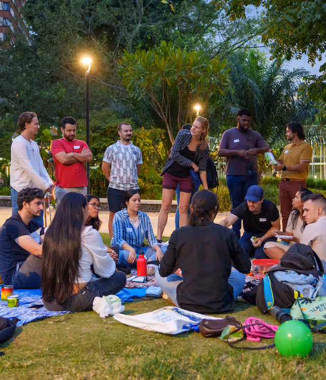 Group of people sitting and standing on grass in a park at dusk, socializing with blankets, bags, and drinks around them.