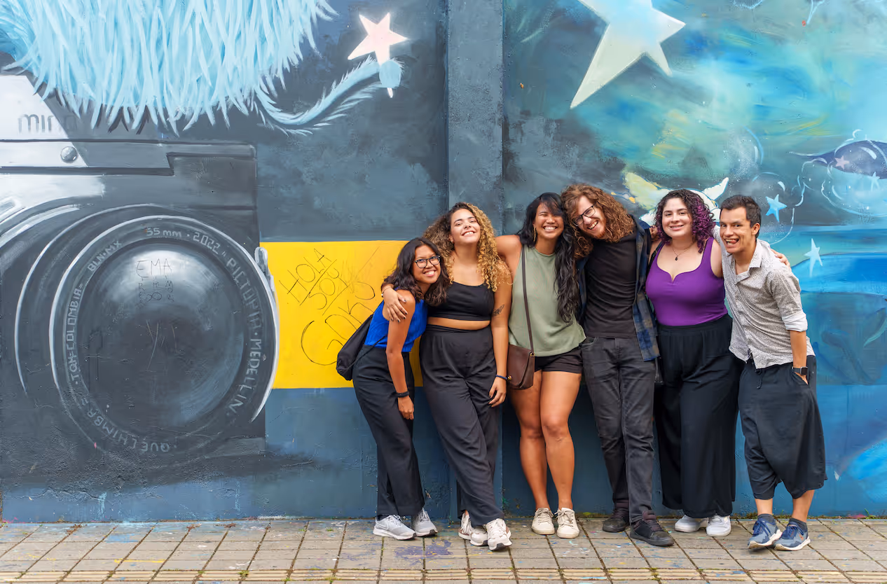 Group of six diverse young adults smiling and posing in front of a colorful mural with a large camera and star designs.