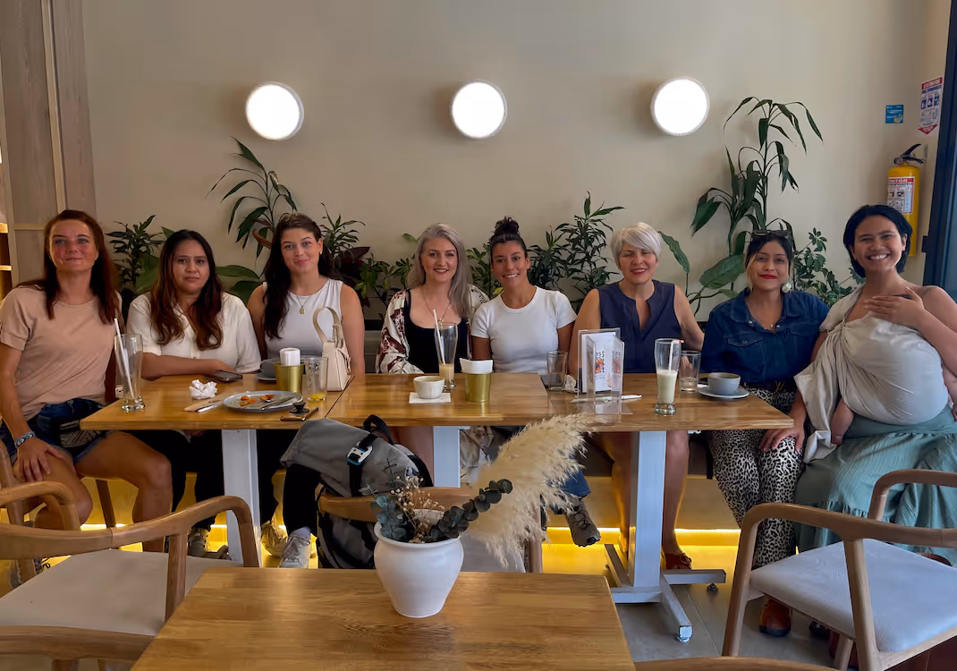 Group of eight women sitting closely together at a wooden table in a cozy café with plants and round wall lights behind them.
