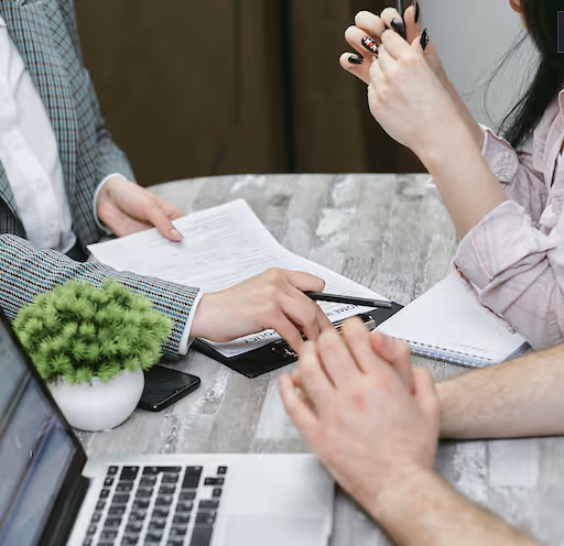Three people discussing documents and notes around a wooden table with a laptop and a small potted plant.