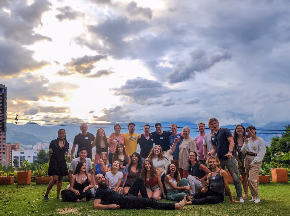 Group of diverse young adults posing and smiling outdoors on green grass with a cloudy sky and cityscape background at sunset.