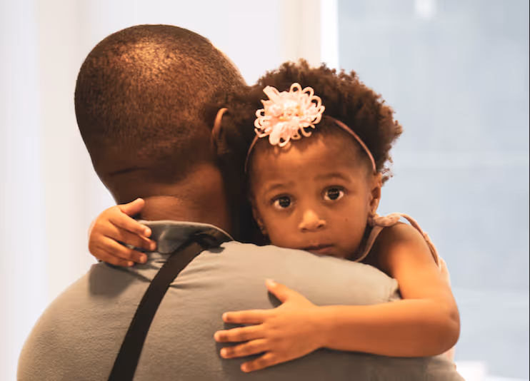 Young girl with a flower headband hugging a man, looking directly at the camera.