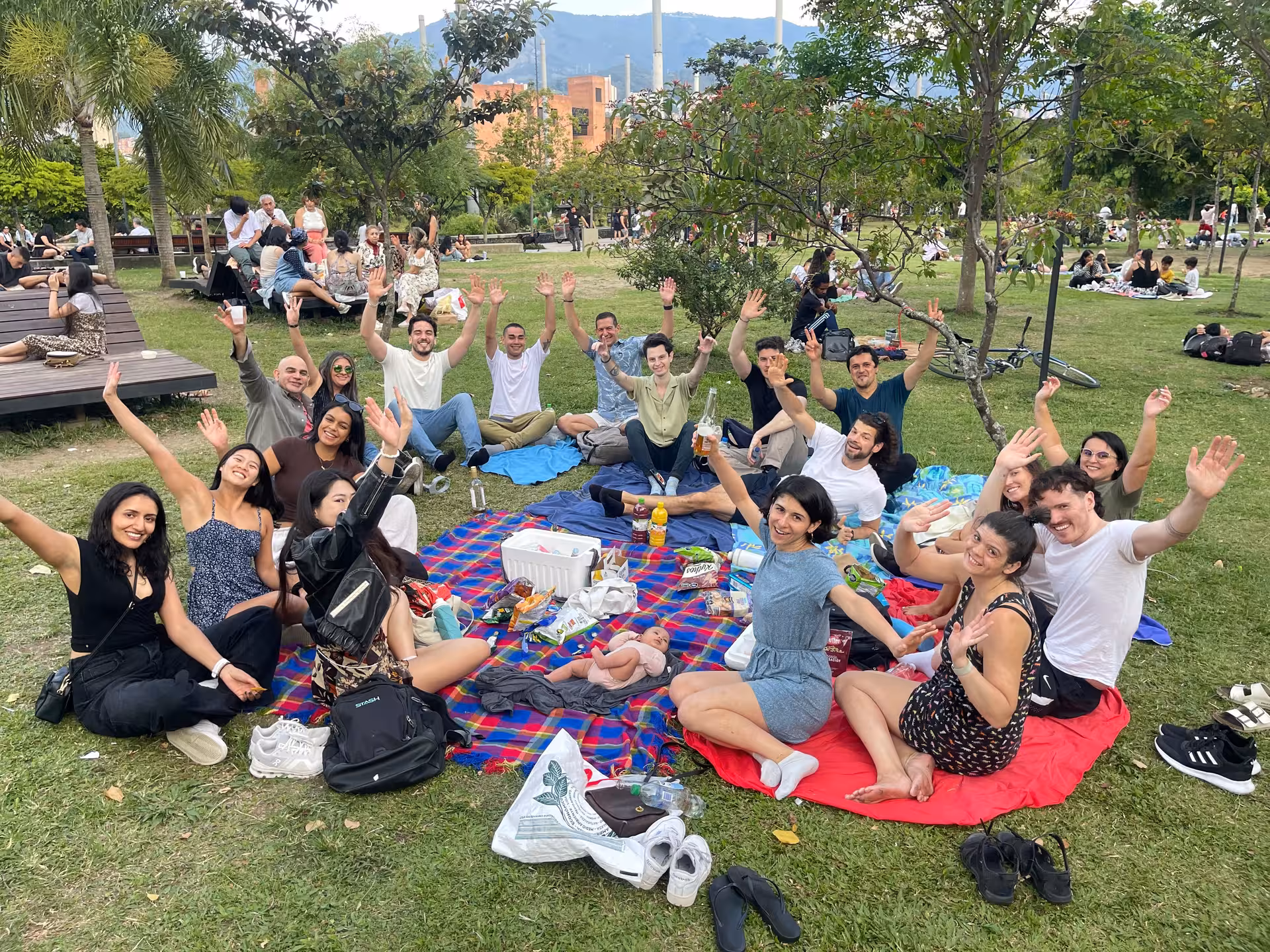 Group of people sitting on blankets having a picnic in a park, smiling and waving at the camera.