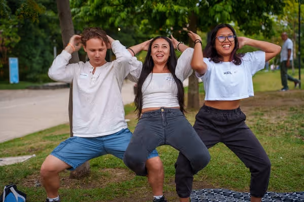 Three young adults outdoors in a park squatting with hands behind their heads and smiling.