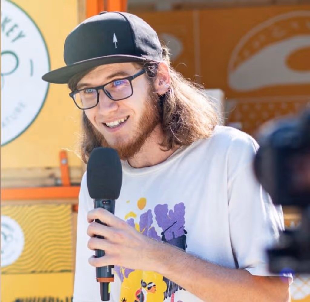 Young man with glasses and a black cap speaking into a microphone outdoors.