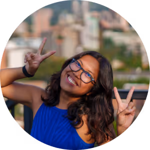 Smiling woman with glasses wearing a blue top, making peace signs with both hands outdoors.