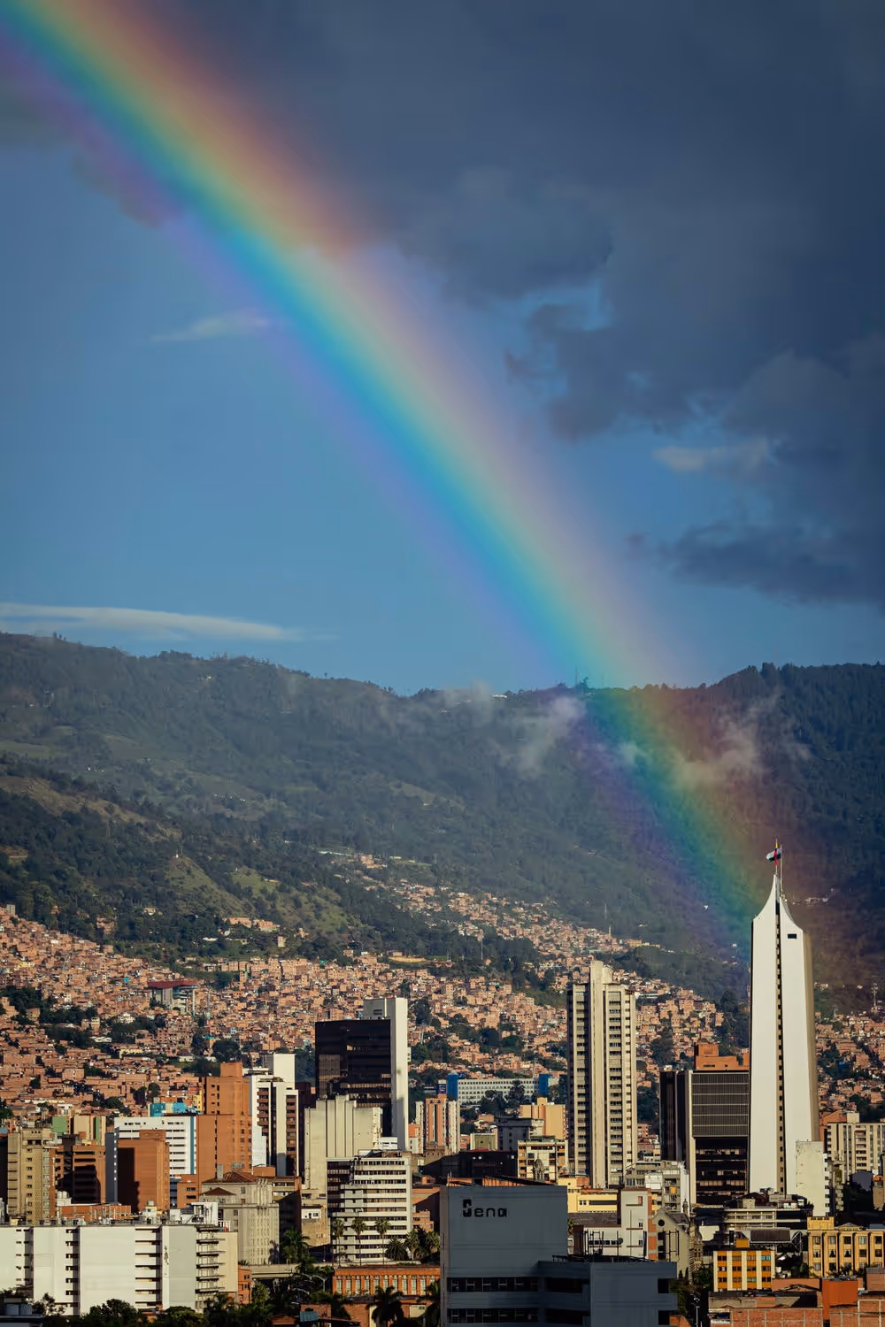 Rainbow arching over a city skyline with tall buildings and green mountains in the background under a partly cloudy sky.