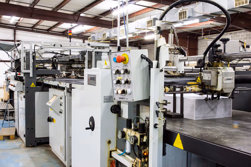 Large industrial printing machine inside a factory with control panel and paper stack.