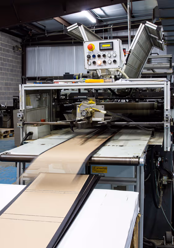 Automated machine in factory cutting cardboard sheets on a conveyor belt.