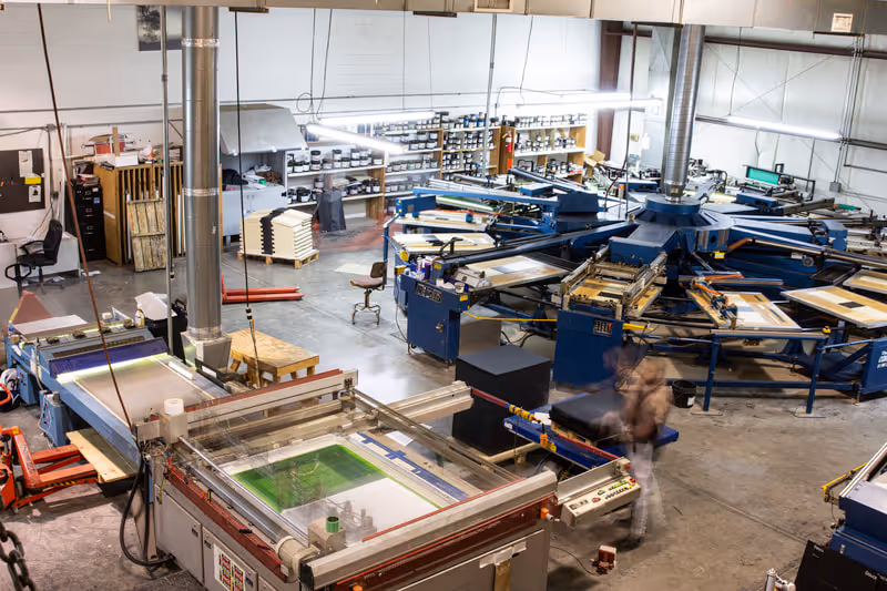 Interior of a large silk screen printing workshop with machines, shelves of inks, and a blurred worker.