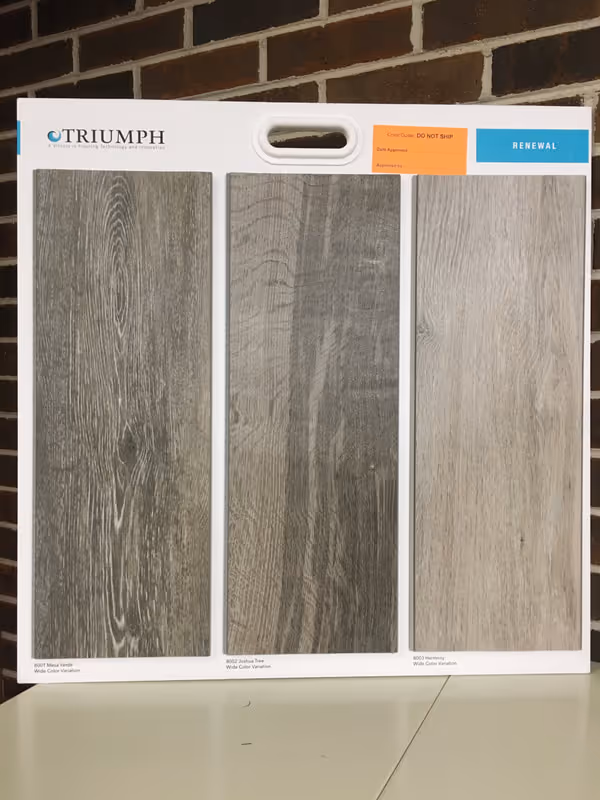 A Triumph brand display board showing three wood flooring samples in gray and beige tones.