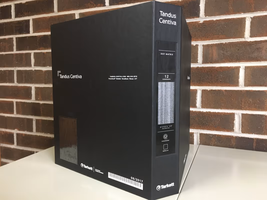 Black sample binder labeled Tandus Centiva with fabric swatch sitting on white table against brick wall.