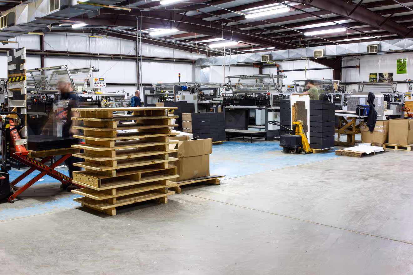 Warehouse with stacked wooden pallets, cardboard boxes, and workers operating machinery.