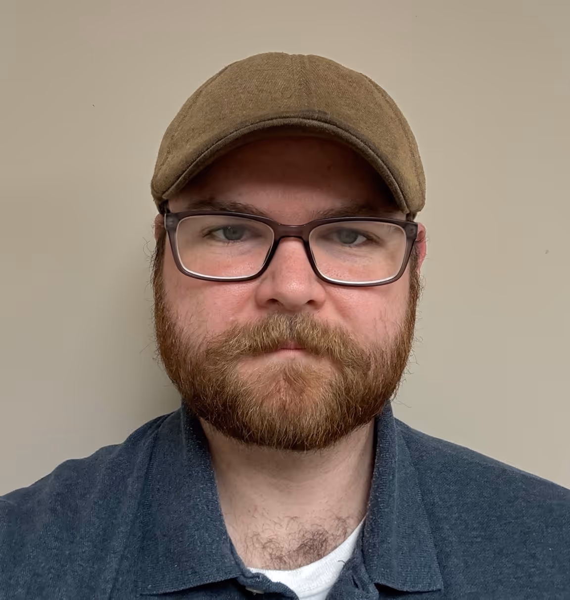 Man with brown beard, glasses, and green cap wearing a dark blue collared shirt with neutral expression.
