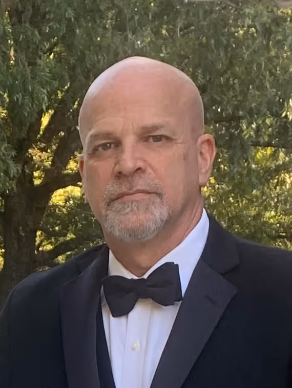 Bald man with a short gray beard wearing a black tuxedo and bow tie outdoors near green trees.