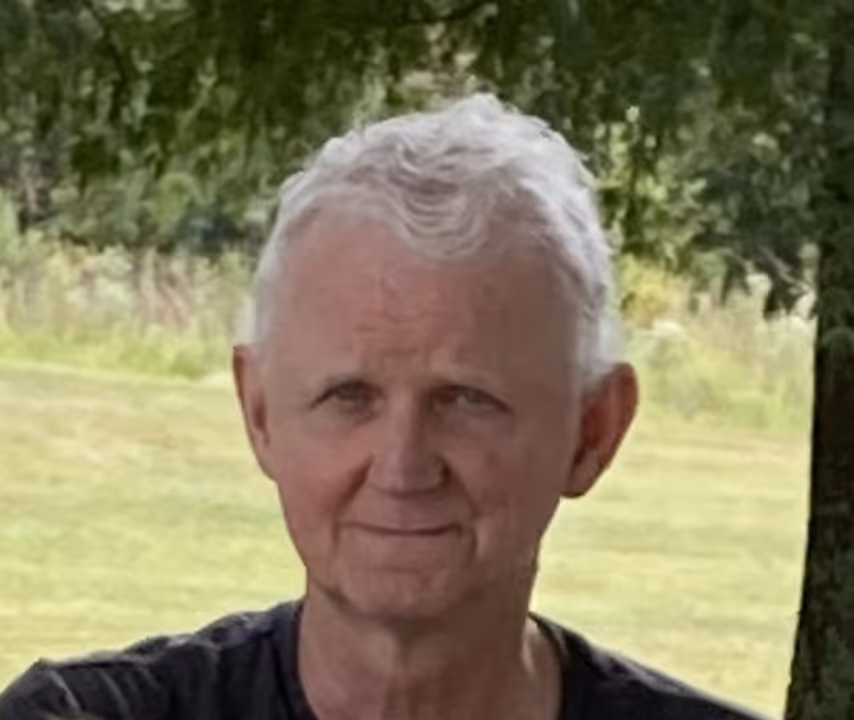 Older man with short white hair sitting outdoors with grass and trees in background.