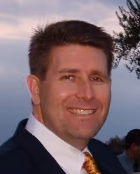 Smiling man with brown hair wearing a dark suit and patterned tie outdoors at dusk.