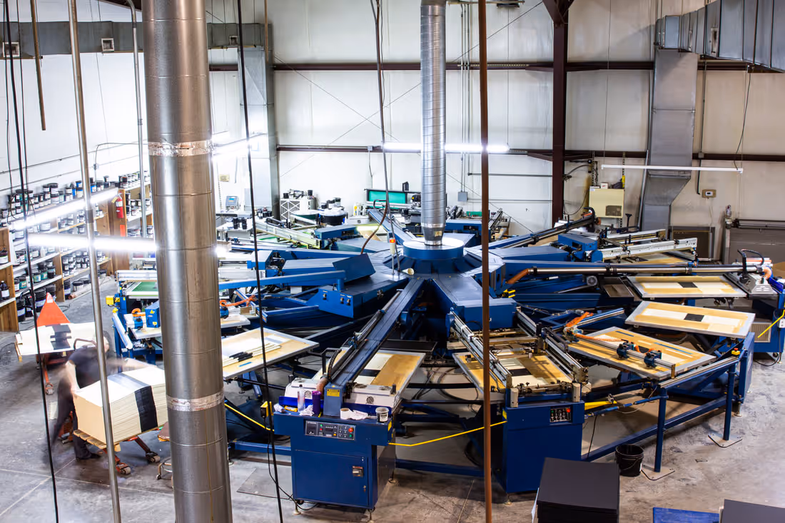 Large industrial screen printing machine in a factory with a worker handling materials nearby.