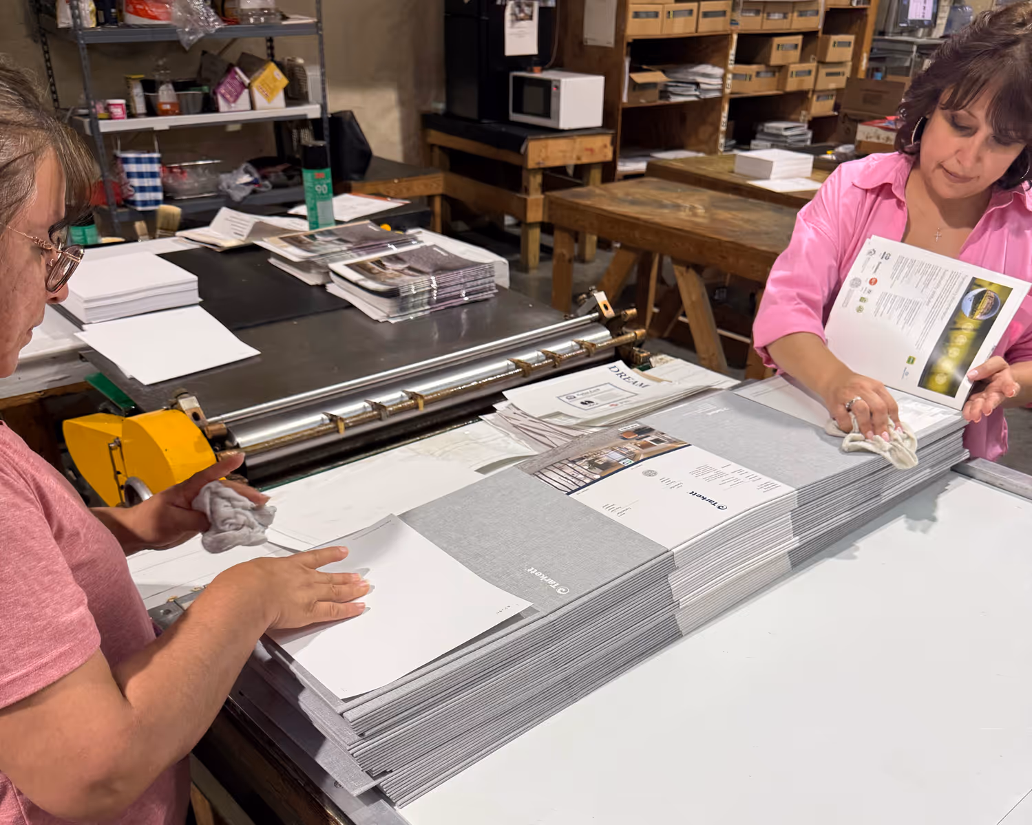 Two women assembling and cleaning stacks of printed booklets in a workshop with shelves and tables