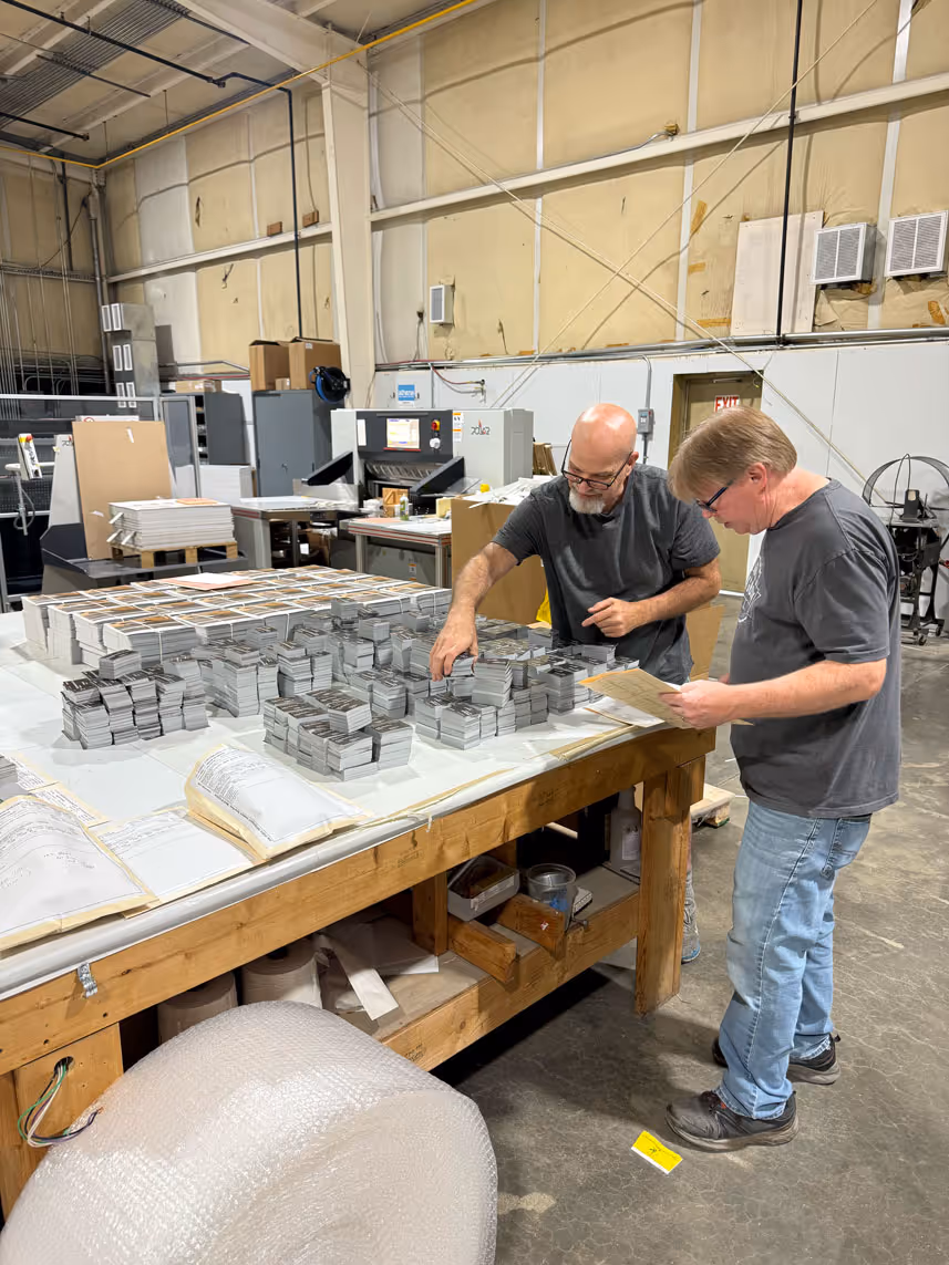 Two men sorting and organizing stacks of printed cards on a large wooden table in a warehouse.