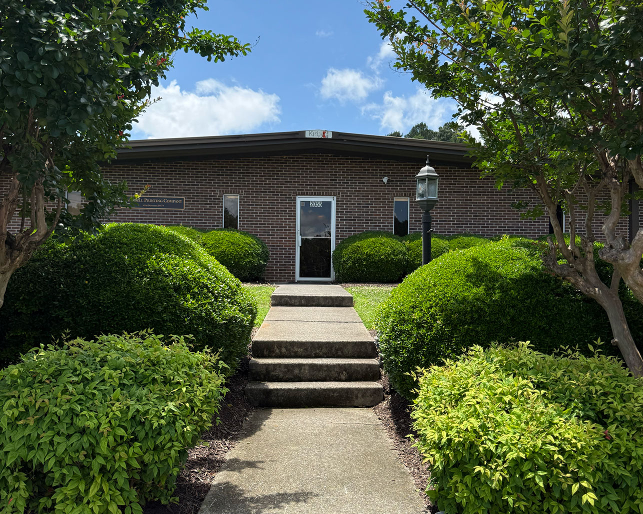 Brick office building entrance with glass door, concrete steps, trimmed bushes, and a lamp post on a sunny day.