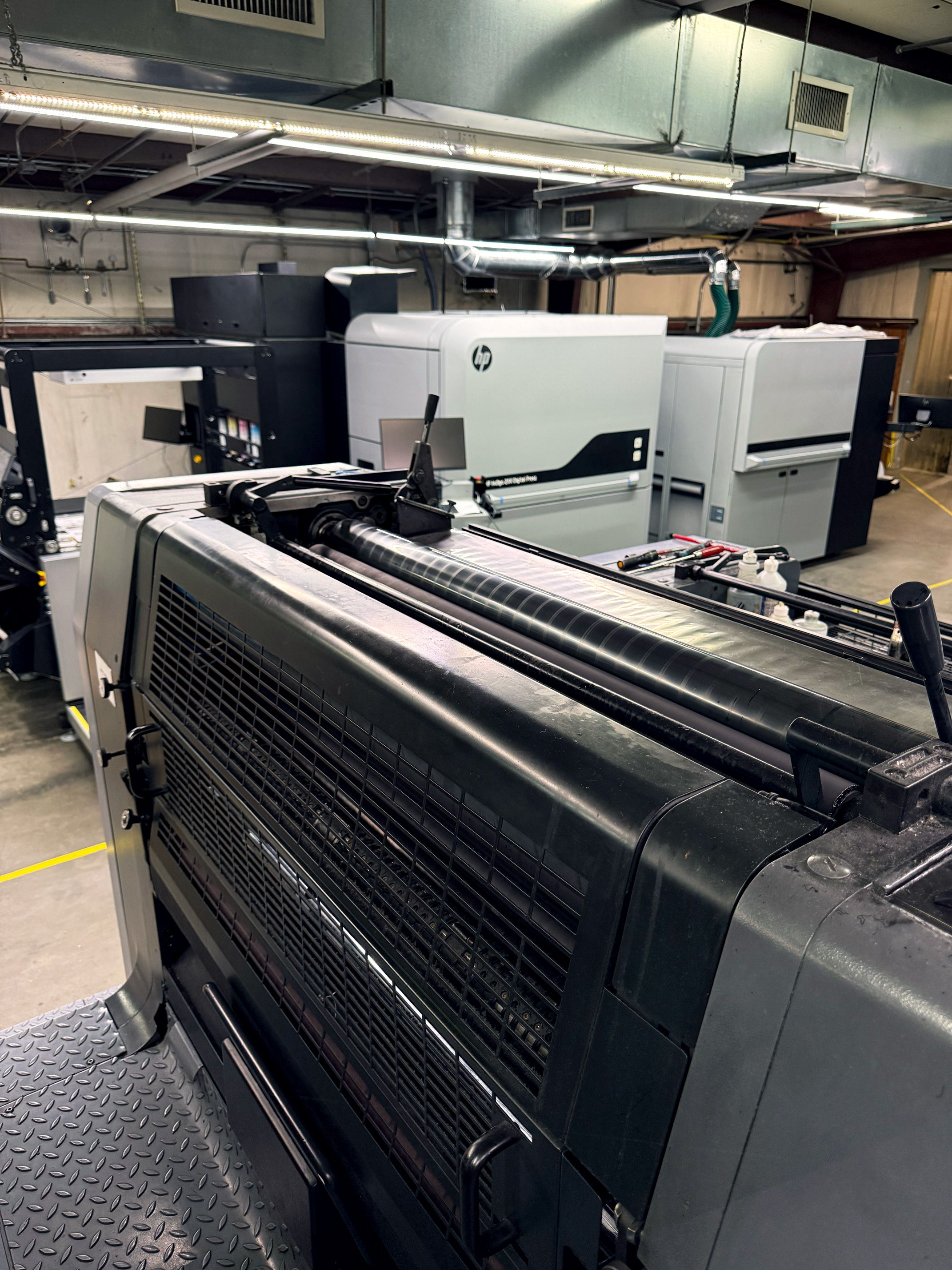 Large industrial printing machines inside a factory room with overhead lights and ductwork.