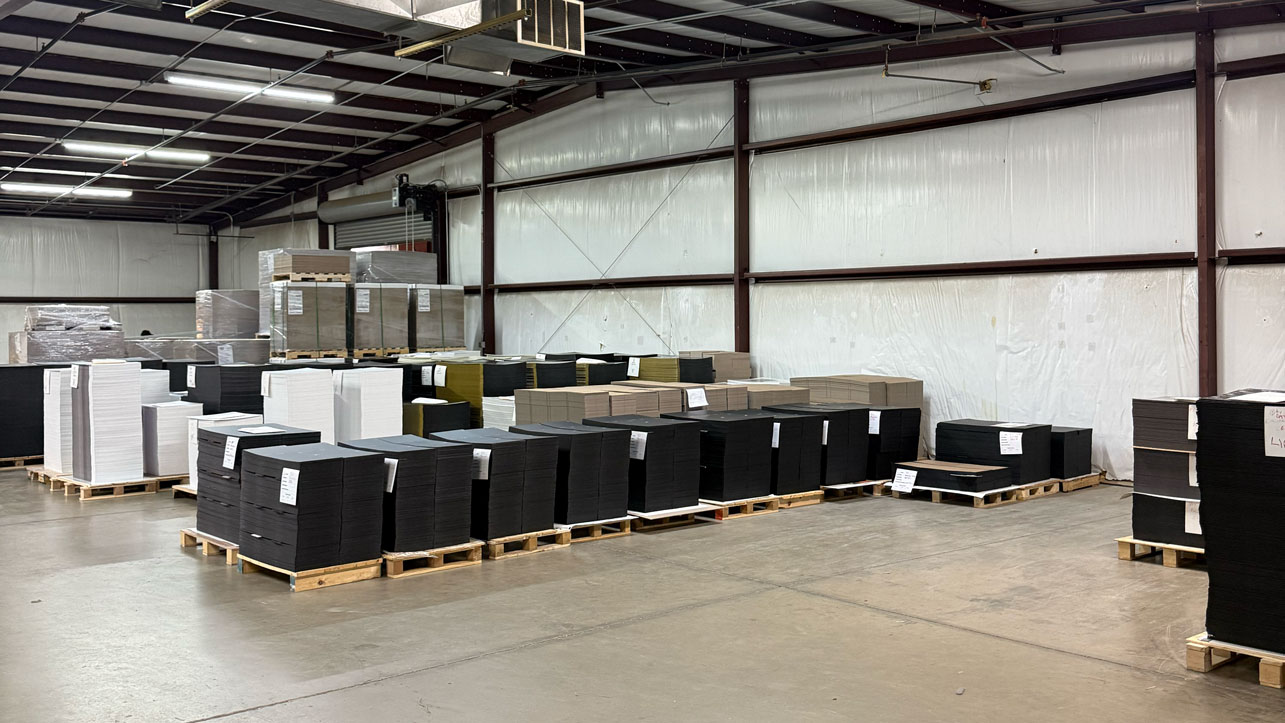 Stacks of black, white, and brown cardboard sheets on wooden pallets inside a warehouse.