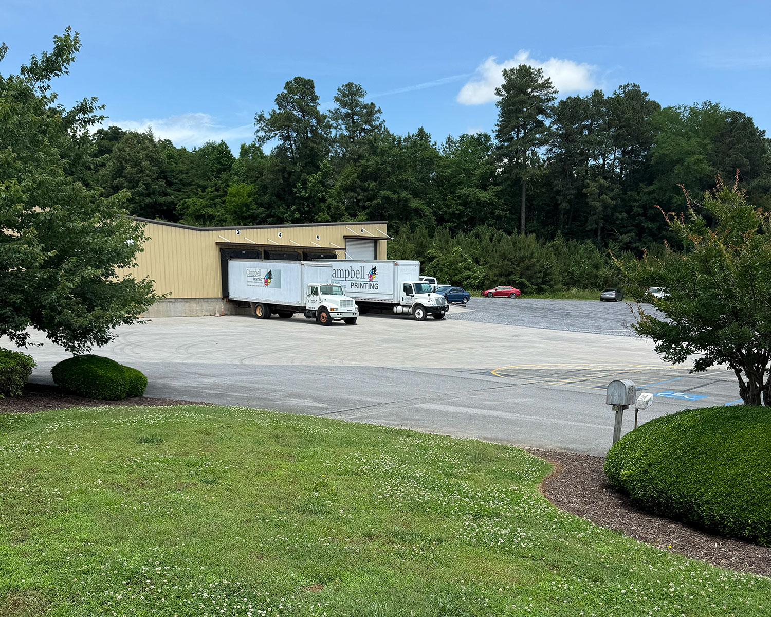 Two Campbell Printing trucks docked at loading bays of a beige warehouse surrounded by trees and cars.