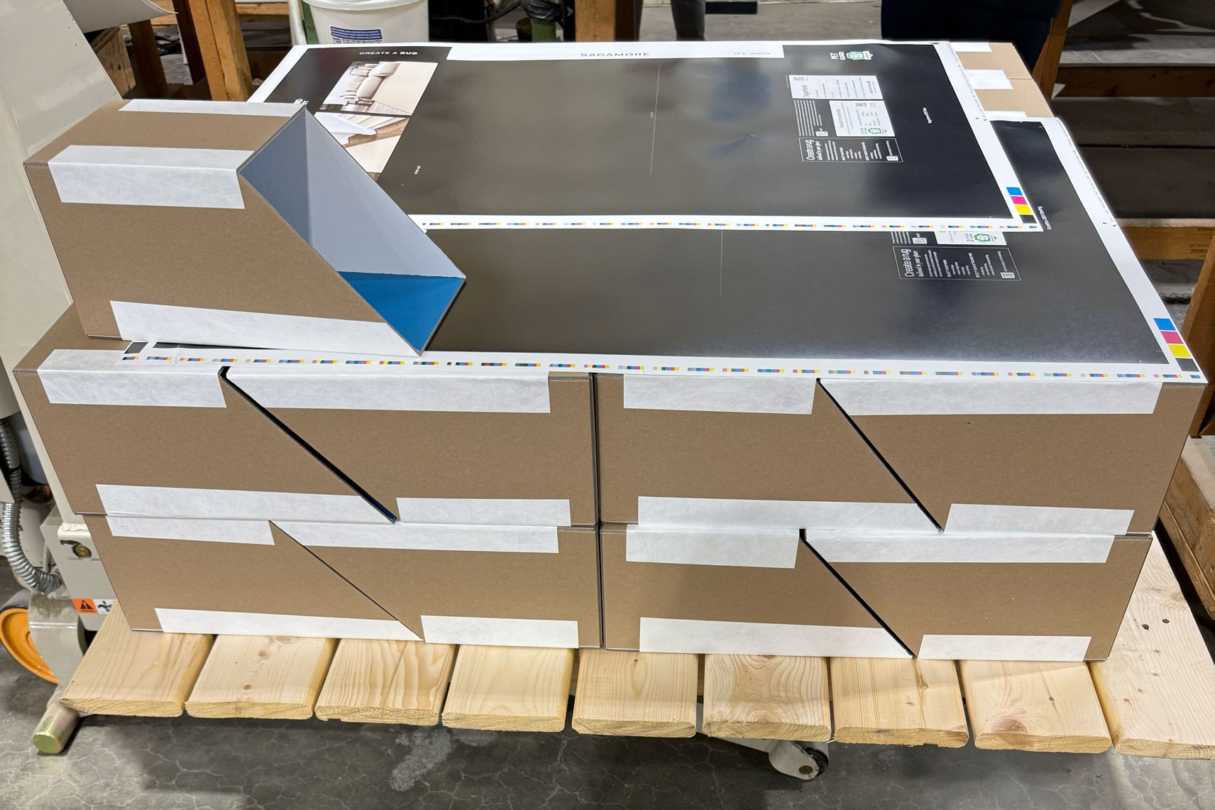 Unassembled brown cardboard box with white tape and print layout resting on wooden pallet in workshop.